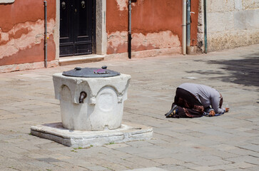 Una mendicante inginocchiata vicino a un pozzo chiede l'elemosina in una piazza di Venezia