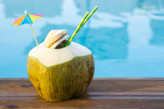 Fresh  green coconut drink with paper straw  and rainbow umbrella  standing on wooden board  near pool water  tropical beach resort background  with copy.  Vacation  exotic resort travel destinations