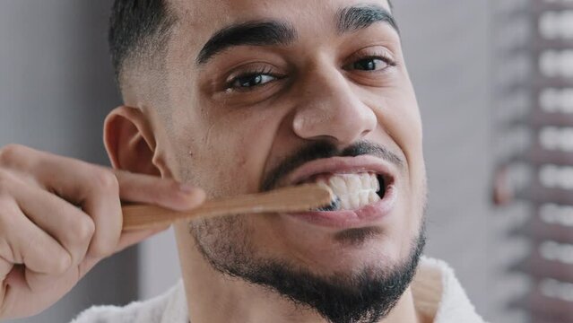 Extreme close up head shot bearded adult handsome Indian Spaniard Arab man brushing teeth in bathroom. Portrait hipster Arabic ethnicity guy doing oral mouth hygienic daily toothy procedure, toothcare