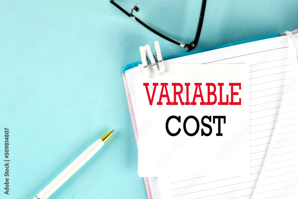 Poster variable cost text on a sticky on notebook with pen and glasses , blue background