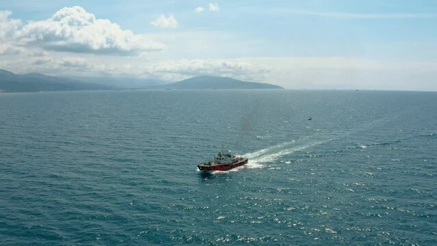 Aerial waterscape with a motor workboat sailing in the cyan quiet sea with its surface shining in the sun. Scenery with sky and hills on the coast in background. Water transportation