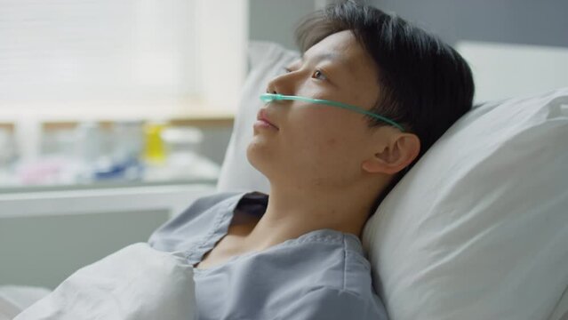 Sick Asian woman with nasal cannula lying on bed in hospital ward and looking away during therapy