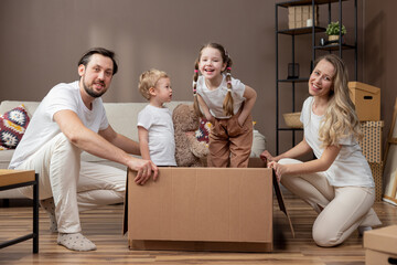 Portrait of a happy young family kneeling parents by big cardboard box in which their children are sitting playing, taking care of daughter and son while moving to a new apartment shared joy.