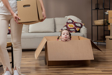 A cheerful little girl in two pigtails hides in a cardboard box left over from the move, in the...