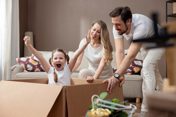 Excited young family having fun on moving day, happy parents pushing cardboard boxes with funny sweet little girl daughter riding in containers, looking at the camera and laughing about moving in.
