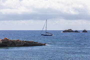 Sailing boat without sails maneuvers between rocks protruding above shallow water under heavy low white clouds. Ibiza, Balearic Islands, Spain