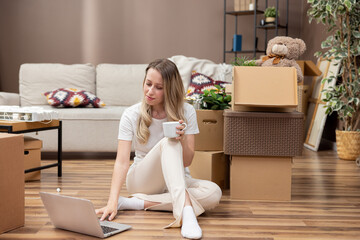 Obraz premium Smiling woman is sitting on floor in middle of new apartment she has moved into, laptop is standing on cardboard boxes, girl is browsing through interior inspirations, looking for furniture to buy.