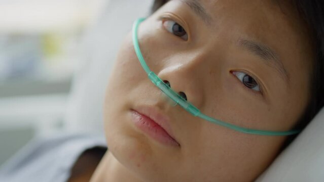 Portrait of sick Asian female patient with nasal cannula lying on hospital bed and looking at camera