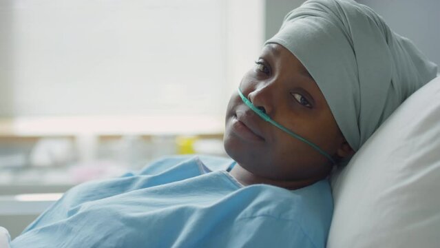 Portrait of sick African American woman with nasal cannula and headwrap lying on bed in hospital ward and posing for camera