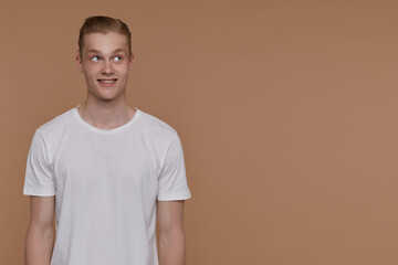 Indoor portrait of young blonde man posing over beige background looking aside at copy space with...
