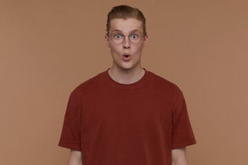 Indoor portrait of young blonde man posing over beige background wears round glasses looking into camera with shocked facial expression