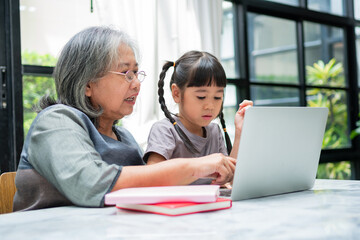 Asian Grandmother with her two grandchildren having fun and playing education games online with a computer notebook at home in the living room. Concept of online education and caring from parents.