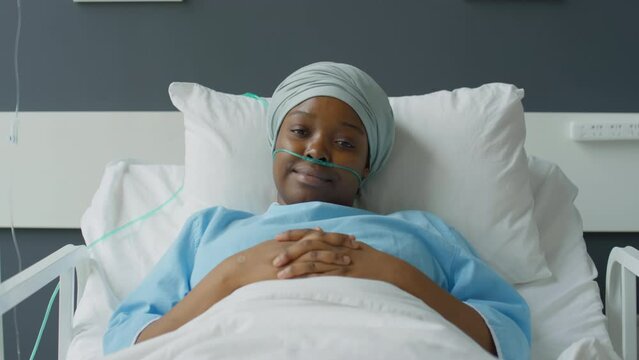 Zoom in shot of young African American female patient with nasal cannula and headwrap lying on hospital bed with hands clasped on her stomach and posing for camera with smile