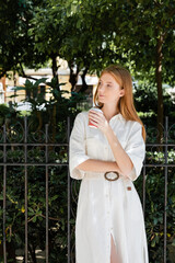 young redhead woman in white dress holding coffee to go near fence in european green park.