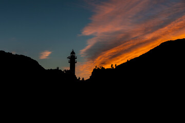 Silhouette of the lighthouse and people on top of the rocks. Beautiful colorful sunset sky. Punta de Teno, near Buenavista del Norte, Santa Cruz de Tenerife.