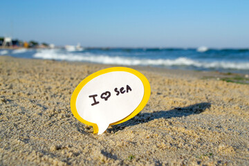 Small stick with paper speech bubble with words I love sea stands on sandy beach near sea water and sea waves with white foam close-up. Concept of travel, tourism, vacation, holiday, relax rest summer