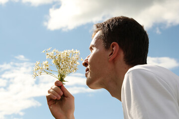 Young man smelling white field flowers in hand and blue sky with white clouds. Photo was taken 5 June 2022 year, MSK time in Russia.
