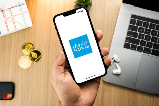 Man holding a smartphone with Charles Schwab financial app on the screen on wooden table with a cryptocurrencies coins and computer. Office environment. Rio de Janeiro, RJ, Brazil. June 2022