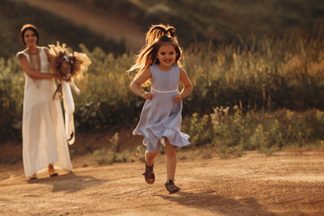 Naklejka na ściany i meble Child girl in a blue dress has fun, laughs and runs away from mom, plays a game in nature