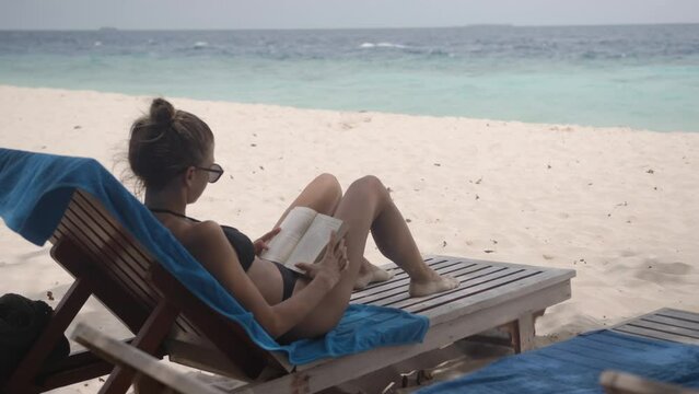 An attractive girl in a swimming suit sits on sun lounger on the beach with a book in her hands. A calm blue sea ahead.
