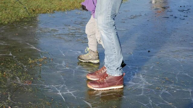Father and son skidding on a frozen pool in a park's grass. Ice has cracks in different directions. Nice warm winter sunlight. Feet on boots doing silly moves having fun.