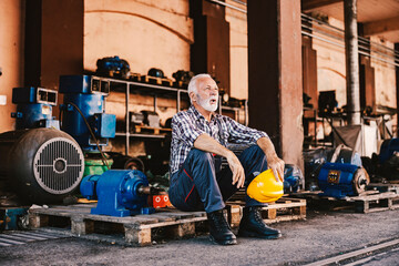 A tired senior factory worker sitting on the pallet at the facility and taking a break after hard...