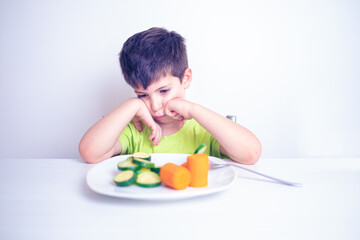 child who hates vegetables useful for his growth looks at the plate with an angry look sitting at the table on isolated white background