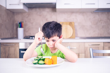 child who hates vegetables useful for his growth looks at the plate with an angry look sitting at the table with the background of the home kitchen