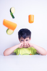 child who hates vegetables useful for his growth is sitting on the table with an angry look and the vegetables fly through the air on a white background