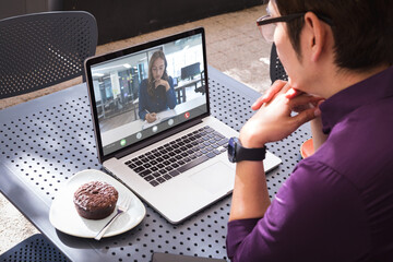 Caucasian businesswoman writing on book during video call with asian male colleague sitting in cafe