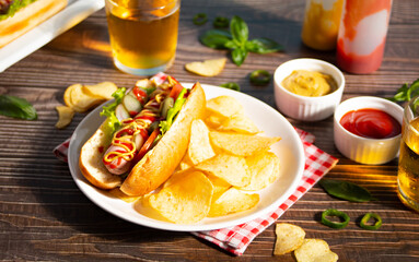 Delicious homemade hotdogs with chips and glasses of beer on wooden table.