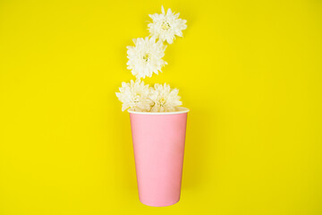 A paper cup for takeaway coffee in pink color lies on a yellow background, white flowers lie out of it. High quality photo
