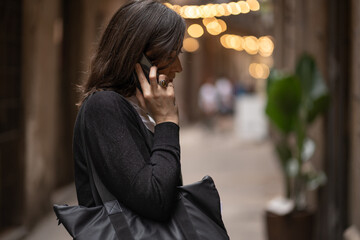 Fototapeta premium Woman talking on the phone on an small old street in Barcelona. Blurred evening city lights in the background.