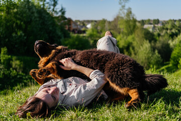 Laughing woman in casual clothes laying on grass and playing with tibetan mastiff dog outdoors - obrazy, fototapety, plakaty