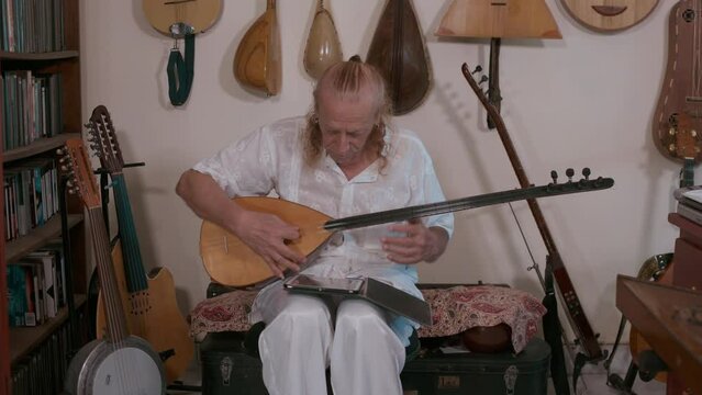 Elderly caucasian man tunes stringed instrument saz in his home studio. Behind him collection of oriental stringed musical instruments.
