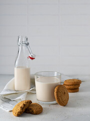 Oatmeal cookies and a glass of melted milk on a light table. A bottle of milk on a light napkin in the background. Light brick background, space for text