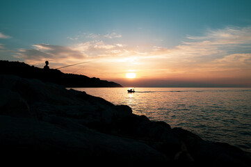 Fototapeta premium SILHOUETTE OF FISHERMEN AT SUNSET ON THE CLIFFS BY THE SEA WITH BOAT IN THE BACKGROUND