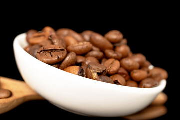 Roasted aromatic coffee beans in a ceramic white saucer with a wooden spoon, close-up, isolated on a black background.