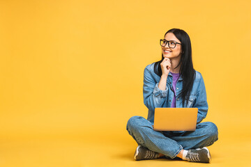 Business concept. Portrait of happy young woman in casual sitting on floor in lotus pose and holding laptop isolated over yellow background. - obrazy, fototapety, plakaty