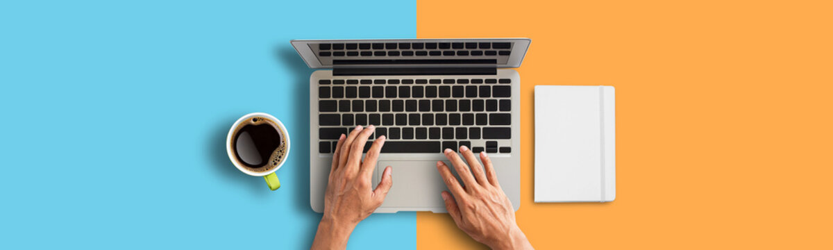 Person hand using laptop computer from above with coffee and smartphone on orange and blue background.