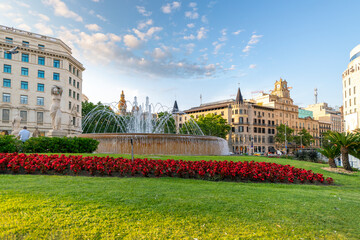 Fountain with red flowers in the foreground at the Plaza de Catalunya in Barcelona , Spain as the sun begins to set in the distance and the city is in view. © Kirk Fisher