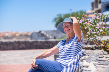 Portrait of cheerful elderly woman dressed in blue sitting outdoors holding hat looking away