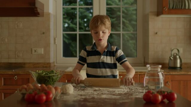 Kid rolling dough standing in the kitchen. Blonde boy baking pizza at home.