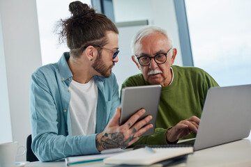 Young educator showing senior student how to solve a problem on project on his tablet while the student typing on a laptop.