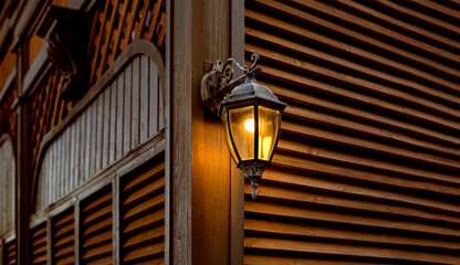 Fototapeta na wymiar iron retro lantern with a glass shade and electric light bulb, object mounted on wooden wall and shutters made of planks, architecture of evening lighting with a warm glow.