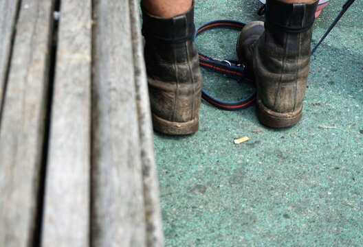 Graues altes Springerstiefelpaar von Mann mit nackten Beinen und Hundeleine auf Platz neben grauer Holzbank in Stadt im Fr&uuml;hling 