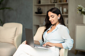 Woman sitting on chair indoors at home apartment, using laptop computer, self employed remote work and small business freelancer, education and leisure time, online shopping, communication technology