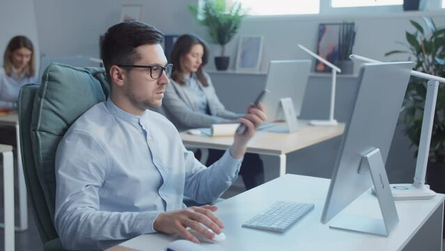 The young attractive businessman is talking on the phone in the office using the computer. Working concept, business concept. People around
