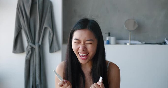 Delighted young Asian pretty woman holding pregnancy test strip, waiting for results standing in the bathroom and rejoicing after great result. Ovulation, fertility, gynecological medical concept