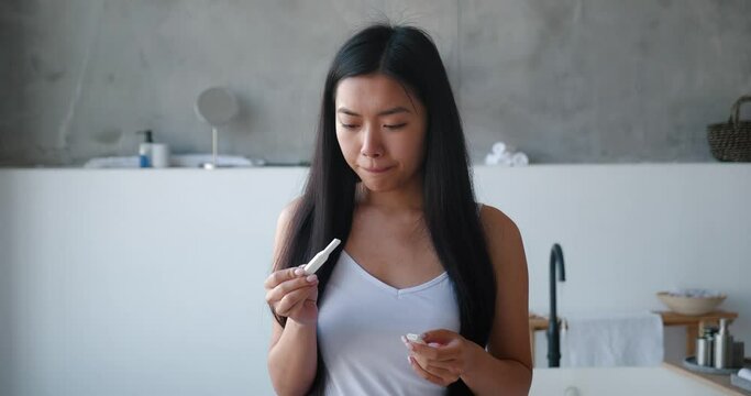 Beautiful delighted young Asian woman holding pregnancy test strip, waiting for results standing in the bathroom and rejoicing after great result. Ovulation, fertility, gynecological medical concept